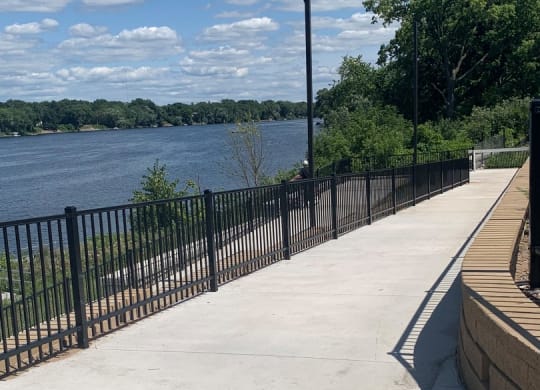 a sidewalk with a bench and fence on the other side of a body of water