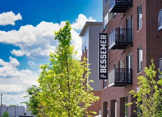 A red brick building with a balcony and a tree in front of it at The Bessemer at Seward Commons Apartments, Minneapolis