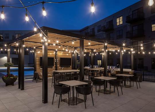A patio with tables and chairs is illuminated by string lights.