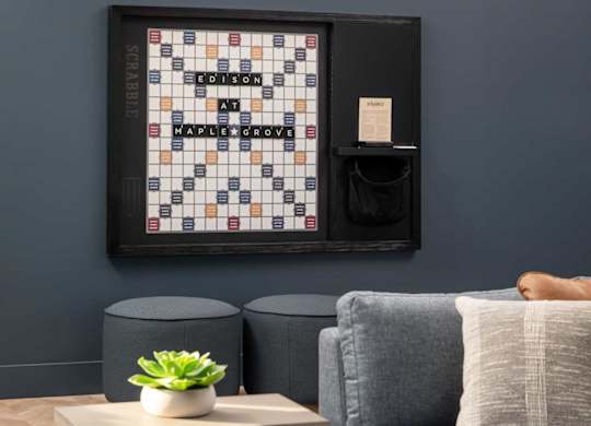 A living room with a grey couch, a small table with a plant and a book, and a framed board game on the wall.at The Edison at Maple Grove, Minnesota, 55311