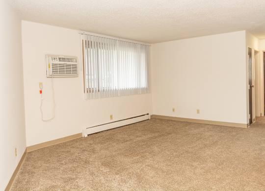 A room with a carpet floor, a window with blinds, and a wall-mounted air conditioner unit.