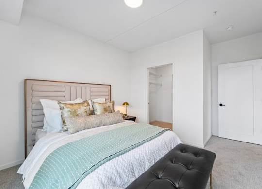 A bedroom with a bed, a black ottoman, and a white door.