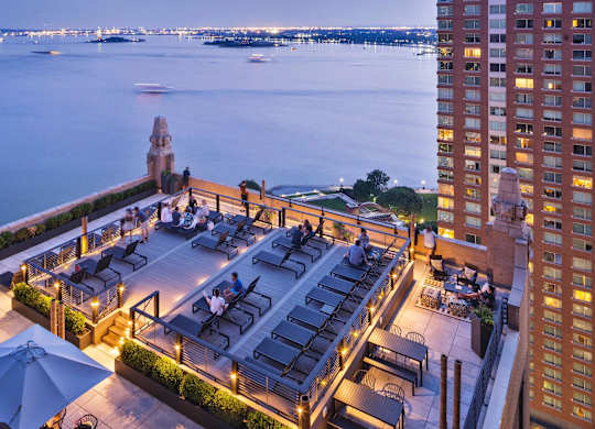 A rooftop bar overlooks a body of water with boats and a city skyline in the distance. at One West, New York, New York