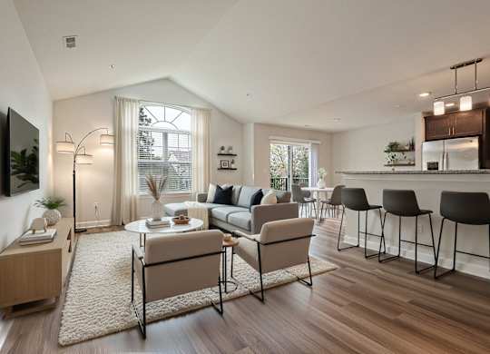 A modern living room with a large window, a dining table set for two, and a kitchen area in the background.
