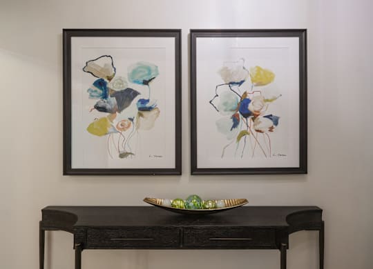Two framed artworks hang on a wall above a dark desk with a green bowl on it.