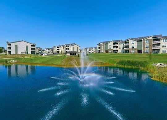 A fountain in the middle of a lake in front of apartment buildings.