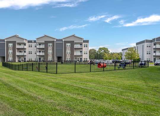 A row of apartment buildings with a grassy field in front and cars parked in the background.