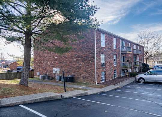 A parking lot in front of a brick building with a tree on the left.