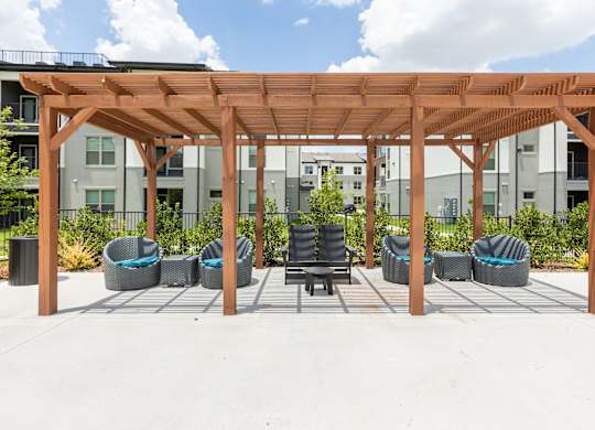 A wooden pergola with wicker chairs and a table in front of apartment buildings.
