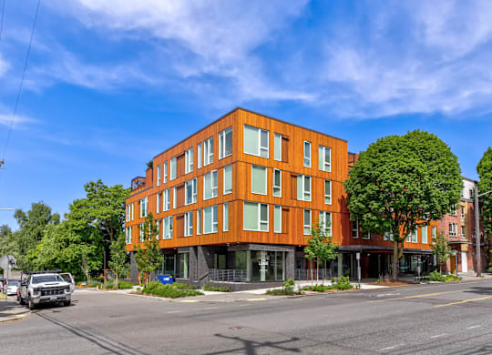 A modern orange building with a flat roof and large windows is situated on a street with cars and trees.