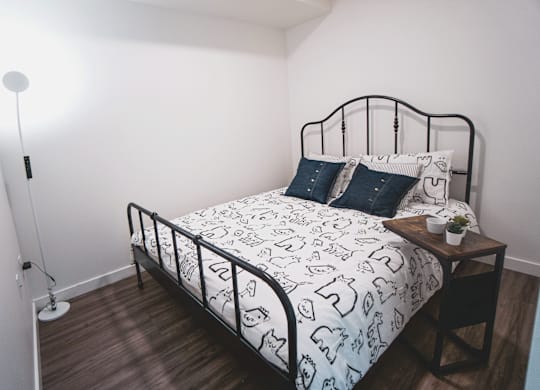 A bed with a white and black patterned comforter sits in a room with a wooden floor and white walls.