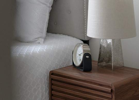 A clock sits on a nightstand next to a bed.