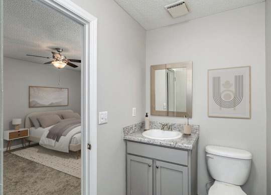 A bathroom with a toilet, sink, and bedroom in the background.