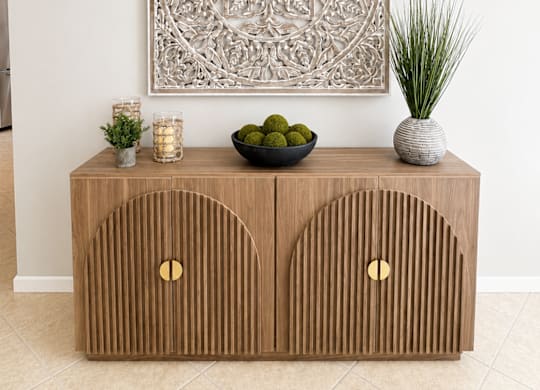 A decorative piece of artwork hangs above a wooden cabinet with two doors, and a bowl of green fruit sits on top of it.
