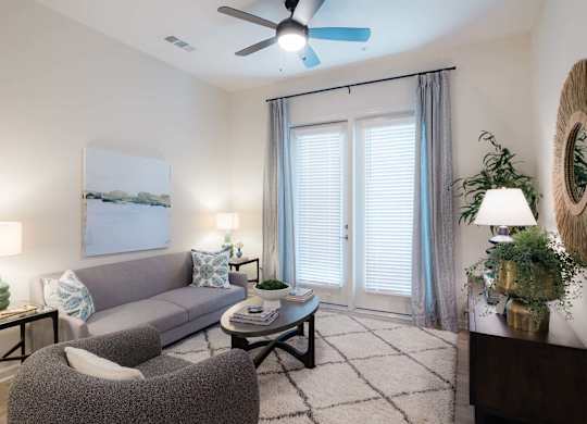 A living room with a grey couch, a grey armchair, a coffee table, a ceiling fan, and a painting on the wall at Hudson at Renaissance Apartments, Wilmington 28405