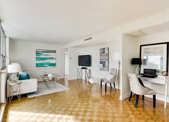 A living room with a white couch, a white chair, a black chair, a white desk, a television, a laptop, a lamp, a painting, and a floor lamp.