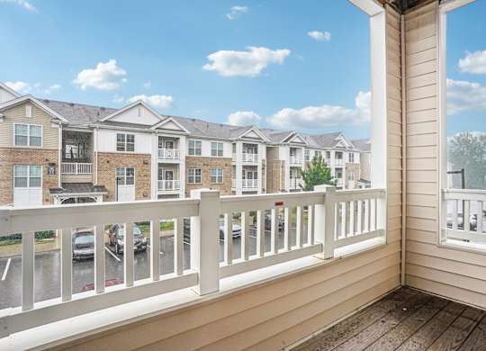 A balcony with a white railing overlooks a parking lot and apartment buildings.
