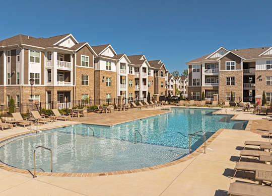 A large swimming pool is surrounded by lounge chairs in front of apartment buildings.