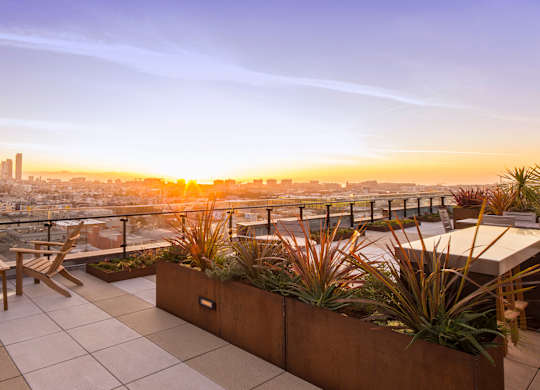 A rooftop patio with a bench and a table with a view of the city at sunset.