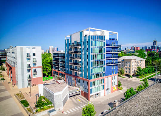 A modern multi-story building with blue and white panels is surrounded by other buildings and greenery.