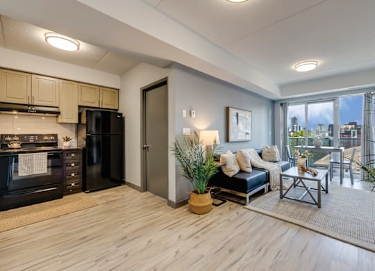 A modern kitchen with a black fridge and microwave, a wooden floor, and a living area with a couch and a coffee table.