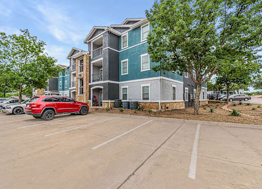 A red car is parked in a parking lot in front of a multi-story building.