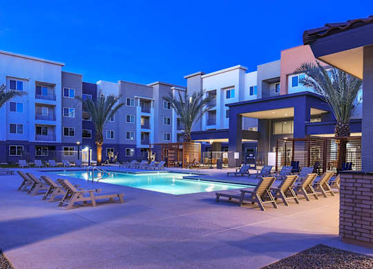 A swimming pool surrounded by lounge chairs and palm trees in front of apartment buildings.