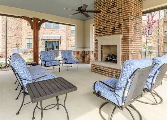 a covered patio with blue chairs and a brick fireplace at Mariposa at Westchester, Grand Prairie, TX