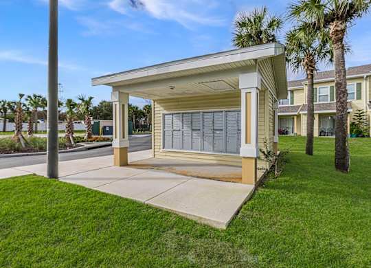 A small building with a porch and a covered entrance sits in front of a grassy area with palm trees.