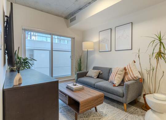 A living room with a grey couch, a wooden coffee table, a white toilet, and a large window.