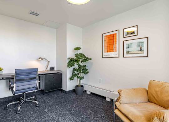 A room with a grey chair, a desk, a potted plant, and two framed pictures on the wall.