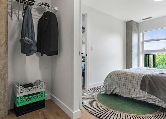 A bedroom with a bed, a coat rack with two coats hanging, and a window with a view of a balcony.