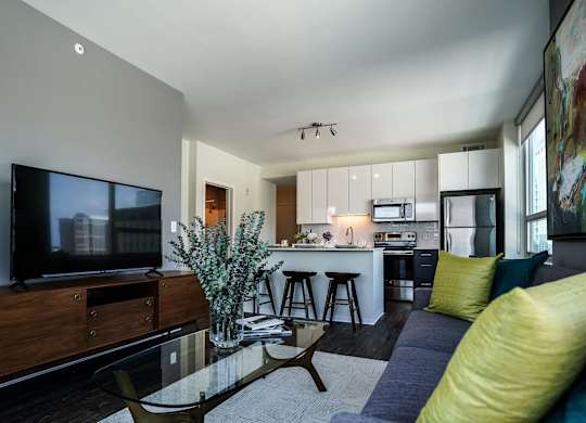 A modern living room with a television, a glass coffee table, and a kitchen in the background.