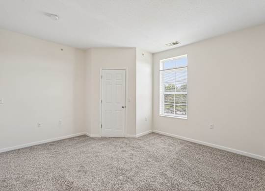 A room with a carpeted floor, a white door, and a window with a view of trees and a building outside.