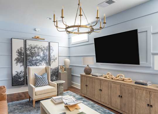 A living room with a white couch, a wooden coffee table, a chandelier, and a large flat screen TV mounted on the wall.
