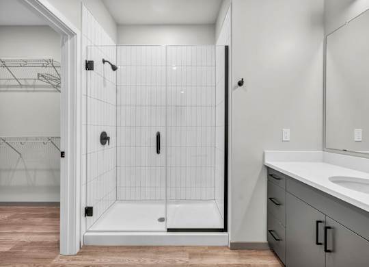 A white tiled shower with a walk-in shower door is in a bathroom with a white sink and a white vanity.