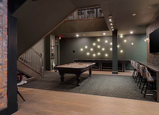 A pool table is in the middle of a room with a brick wall and a staircase.
