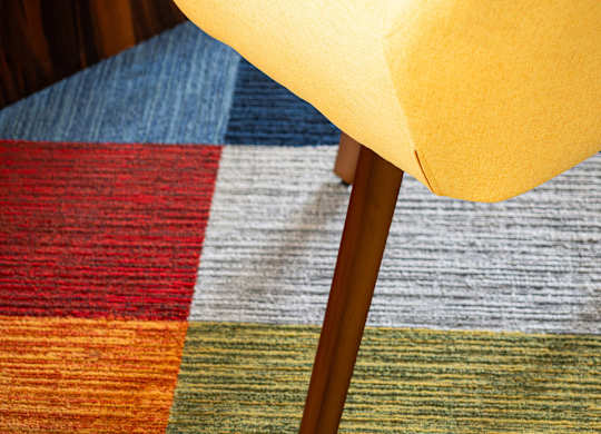 A yellow chair with wooden legs sits on a colorful carpet.