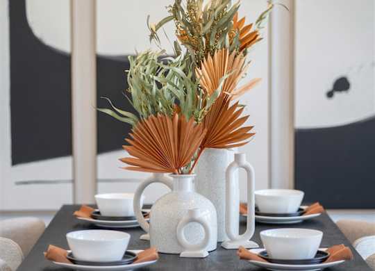 A table with a vase of dried flowers and cups on it.