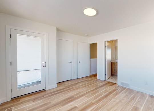 A room with white walls and wooden flooring with a door on the left and a window on the right.