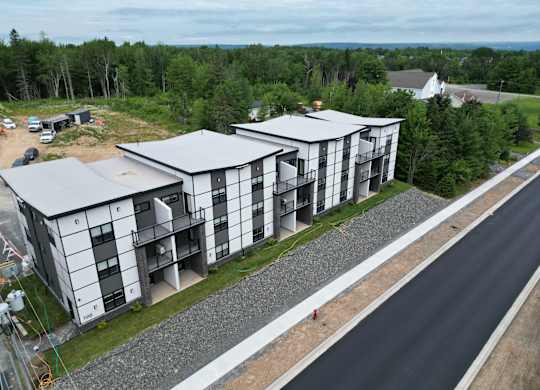 A modern building with a grey and white exterior is surrounded by a gravel area and a paved road.