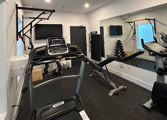 The image shows a room with a black and white interior, featuring a treadmill and a small table with a monitor on it.