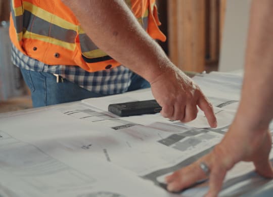 A person in an orange vest is pointing at a blueprint.