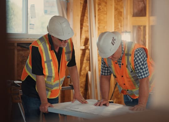 Two construction workers wearing safety helmets and vests are examining a blueprint on a table.