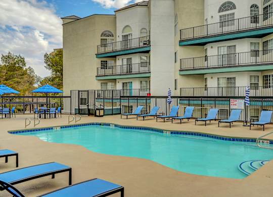 A swimming pool surrounded by blue lounge chairs and umbrellas in front of a multi-story apartment building.
