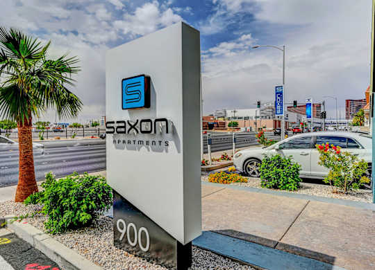 A sign for Saxon Apartments stands in front of a street with a palm tree.