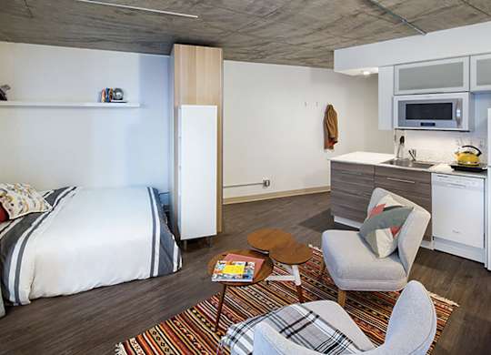 A bedroom with a bed, chair, and a kitchen area.