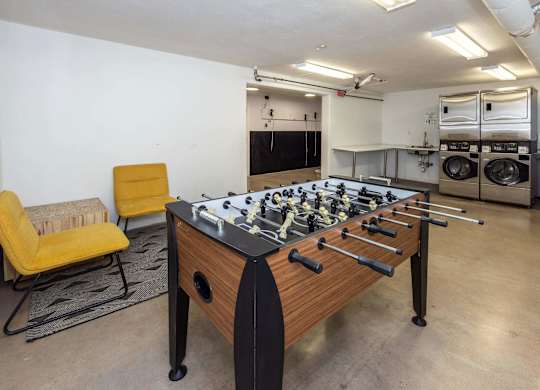 A foosball table is in the middle of a room with a yellow chair and a washing machine in the background.