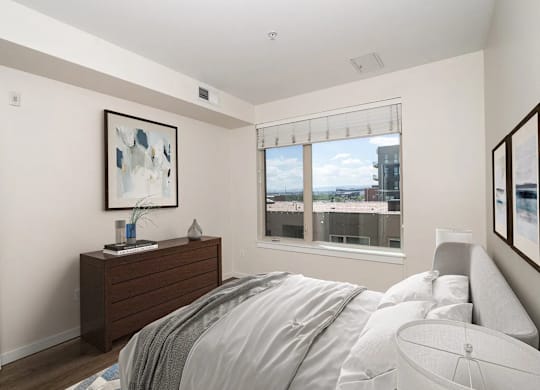 A bedroom with a bed, nightstand, and a window overlooking a cityscape.