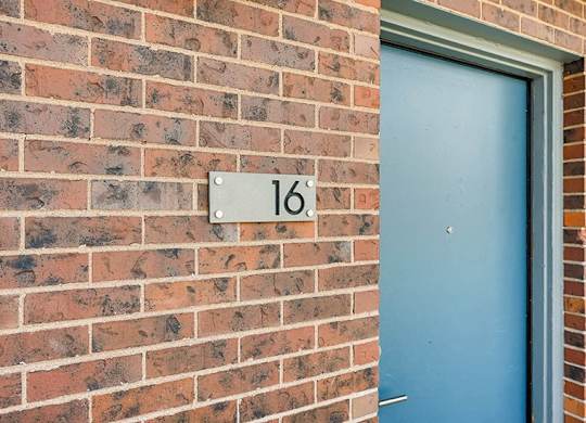 A blue door is on the right side of a brick wall with a white number 16 on a white plaque.
